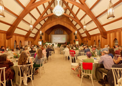 A large group of people sits at round tables in a wood-paneled hall with vaulted ceilings, chandeliers, and a stage at the front showing a presentation. The atmosphere is formal and festive.