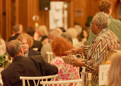 A woman in a patterned dress serves snacks from a basket to guests seated at round tables during a social event in a warmly lit room.