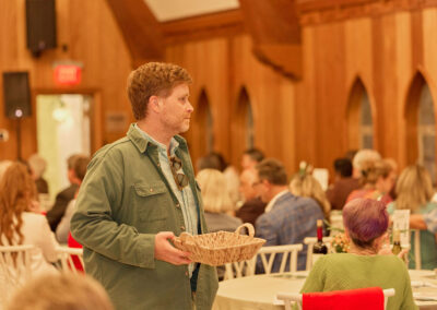 A man in a green jacket holds a woven basket while standing in a room filled with seated people at round tables. The setting has wood-paneled walls and arched windows, suggesting a social or community event.