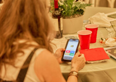 A person with long hair is seated at a table, looking at a smartphone. On the table are red cups, a wine bottle, a plant, and some papers and food. The focus is on the phone and table items.