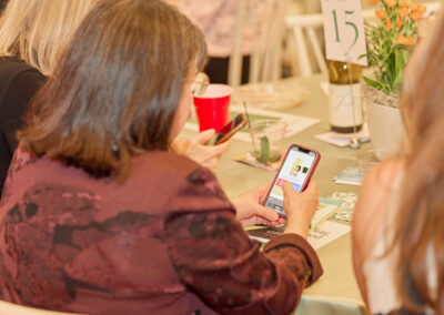 A woman with shoulder-length brown hair and a maroon jacket sits at a table, using her smartphone. Other people are nearby. Table decorations, a red cup, and a sign with the number 15 are visible.