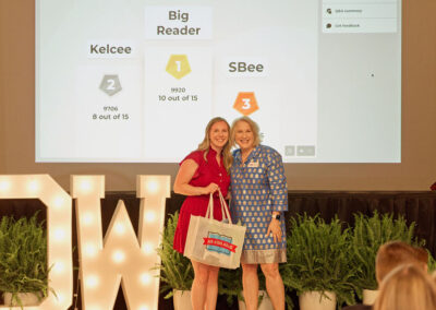 Two women smile and pose together on stage, one holding a gift bag. Behind them is a screen displaying Big Reader rankings for Kelcee and SBee, and large lit-up letters and green plants decorate the stage.