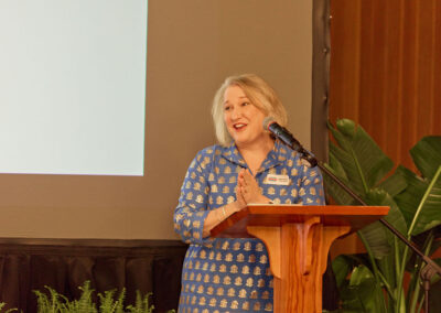 A woman with shoulder-length gray hair, wearing a blue patterned dress, stands at a wooden podium speaking into a microphone. She is smiling, with her hands clasped. Green plants and a large screen are in the background.