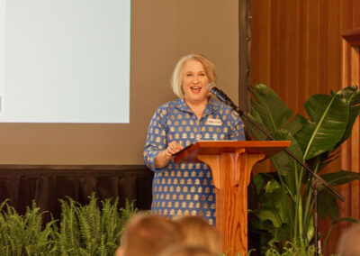A woman with short blonde hair in a blue patterned dress stands at a wooden podium, smiling and speaking into a microphone. She is indoors, with plants nearby and a projection screen behind her.