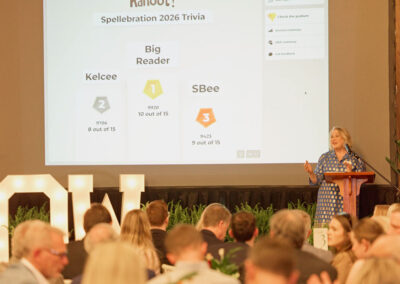 A woman speaks at a podium in front of a projected screen displaying trivia results, as an audience watches. Large lit letters spell OWL and a leaderboard shows scores for Kelcee, Big Reader, and SBee.