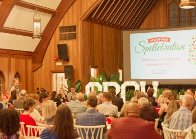 A large group of people sits at tables inside a wooden hall, attending an event. A speaker stands at a podium, and a screen displays “AR Kids Read Spellebration” and “Thank you for investing in Arkansas students!”.