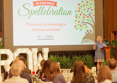 A woman speaks at a podium in front of an audience. Behind her is a screen displaying AR Kids Read Spellebration. Thank you for investing in Arkansas students! and a colorful tree graphic. Large light-up letters spell ROW.