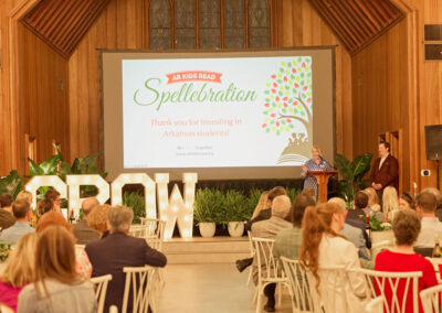 A woman speaks at a podium during the AR Kids Read Spellabration event in a wood-paneled hall. The screen behind her thanks guests for investing in Arkansas students. People sit and listen at decorated tables.