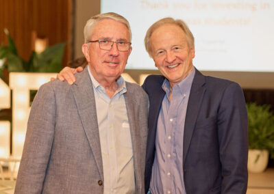 Two older men in suits smiling and standing close together at an indoor event; a blurred projection screen and decorative lights are visible in the background.