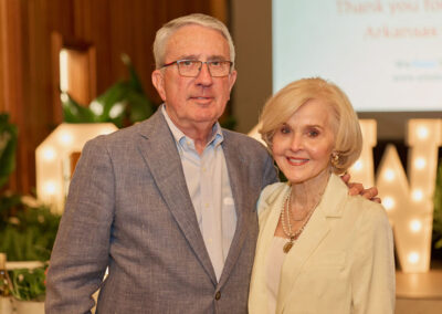 An older man in a blue blazer and glasses stands next to an older woman in a cream jacket, both smiling at an indoor event with plants and decorative lights in the background.