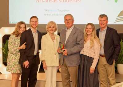 Six adults stand together indoors, smiling at the camera. The man in the center holds an award. They are dressed in business and semi-formal attire. A presentation screen with text is visible in the background.