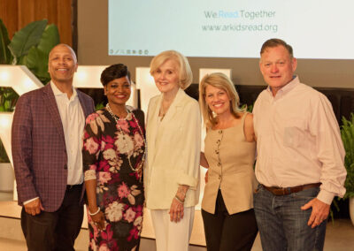 Five adults, three women and two men, stand together smiling in a well-lit indoor setting with greenery and a screen showing WeRead Together and www.arkidsread.org in the background.