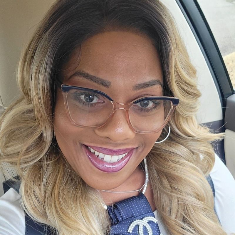 Smiling woman with wavy blonde hair, wearing large glasses, a white top, and a necklace with a Chanel logo, sitting in a car.