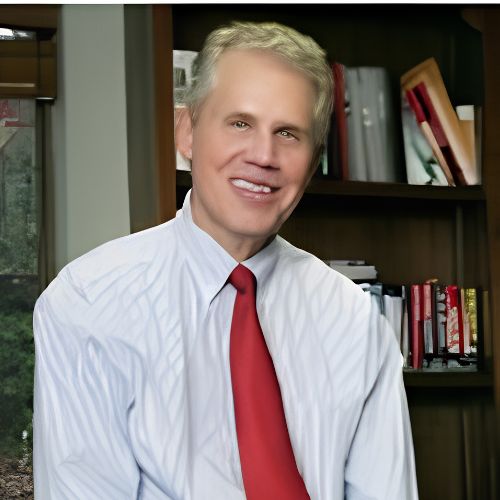 A smiling man in a white dress shirt and red tie sits in front of a bookshelf filled with books in an office setting. Greenery is visible through a window to the left.