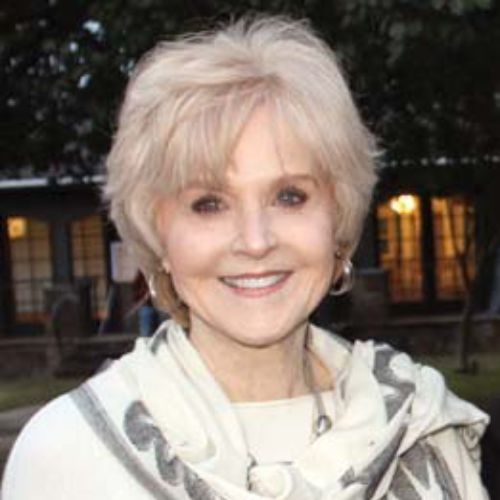 A smiling older woman with short, light blonde hair is wearing a light-colored scarf and standing outdoors, with trees and a building with lit windows in the background.