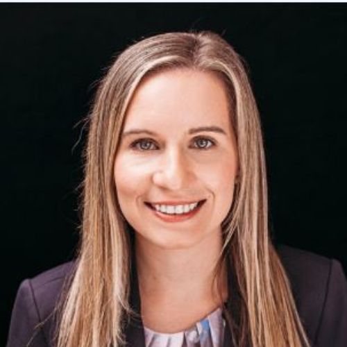 A woman with straight, long blonde hair smiles at the camera. She is wearing a dark blazer over a patterned top with a black background behind her.